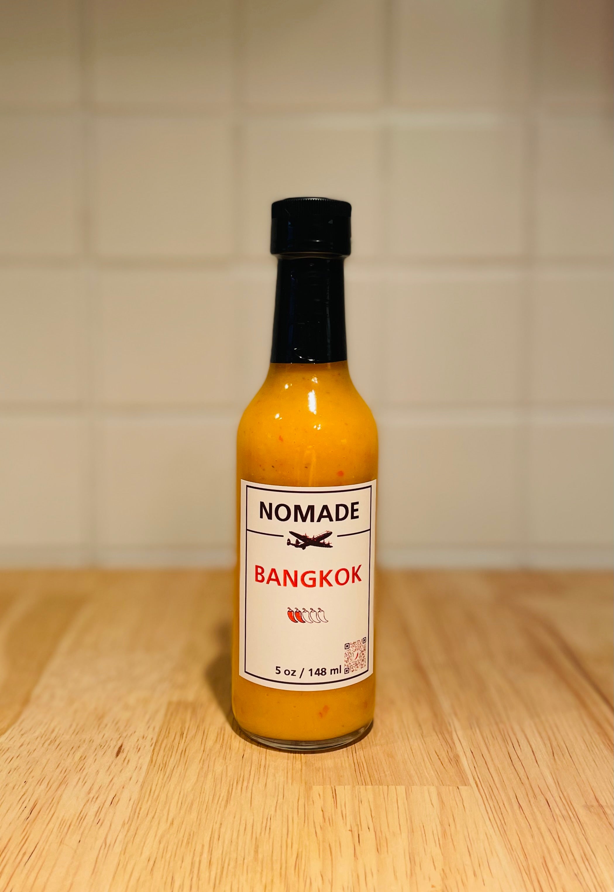 Bottle of Nomade Bangkok hot sauce on a wooden surface with a tiled wall background