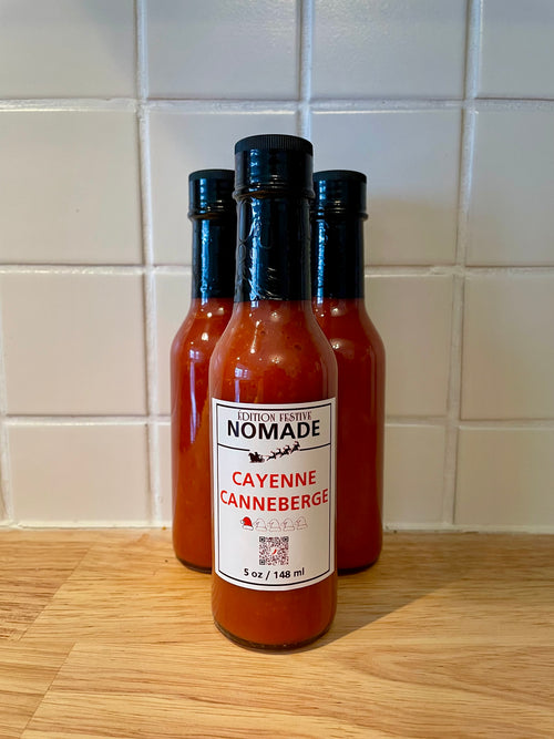 Three bottles of Nomade Cayenne Canneberge festive hot sauce on a wooden surface with a tiled wall background.