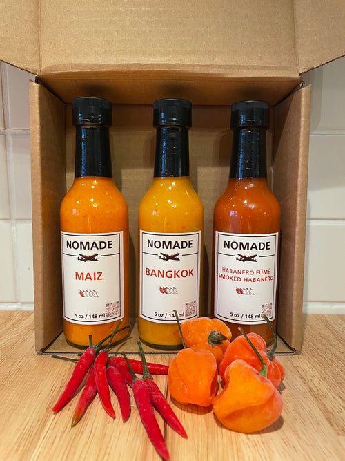 Three bottles of Nomade hot sauce in a cardboard box with peppers on a wooden surface.