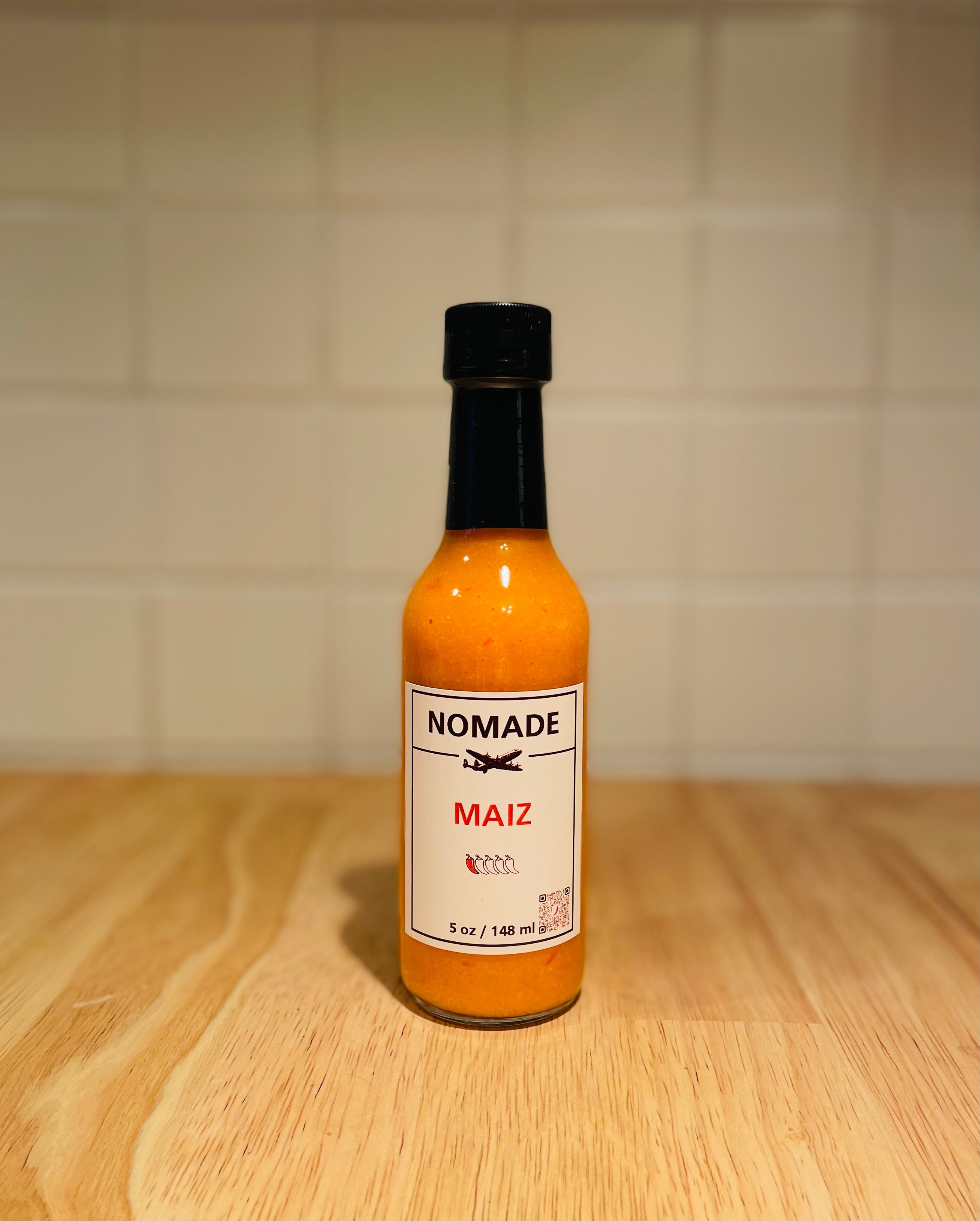 Bottle of Nomade Maiz hot sauce on a wooden surface with a tiled wall background