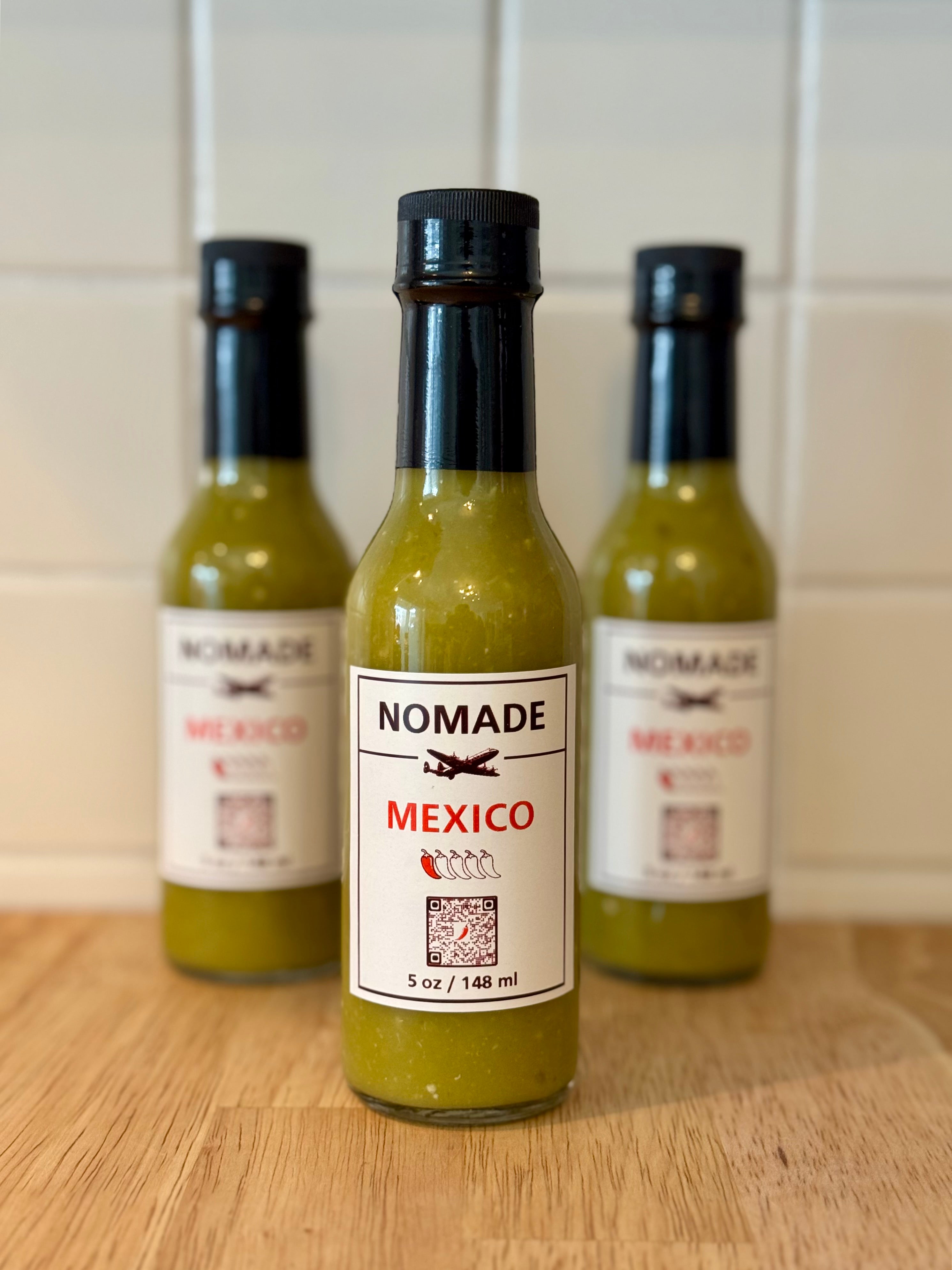 Three bottles of Nomade Mexico hot sauce on a wooden surface with a tiled wall background.