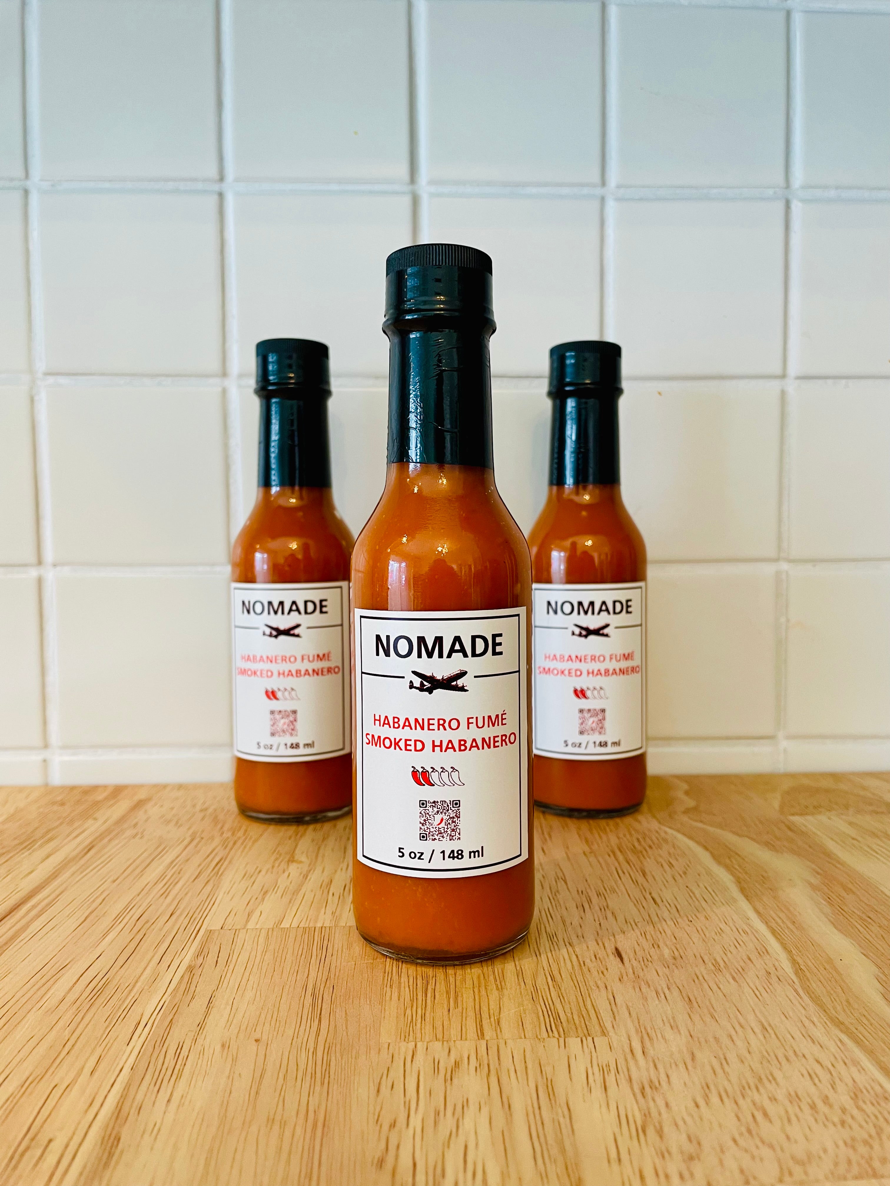 Three bottles of Nomade hot sauce on a wooden surface with a tiled wall background