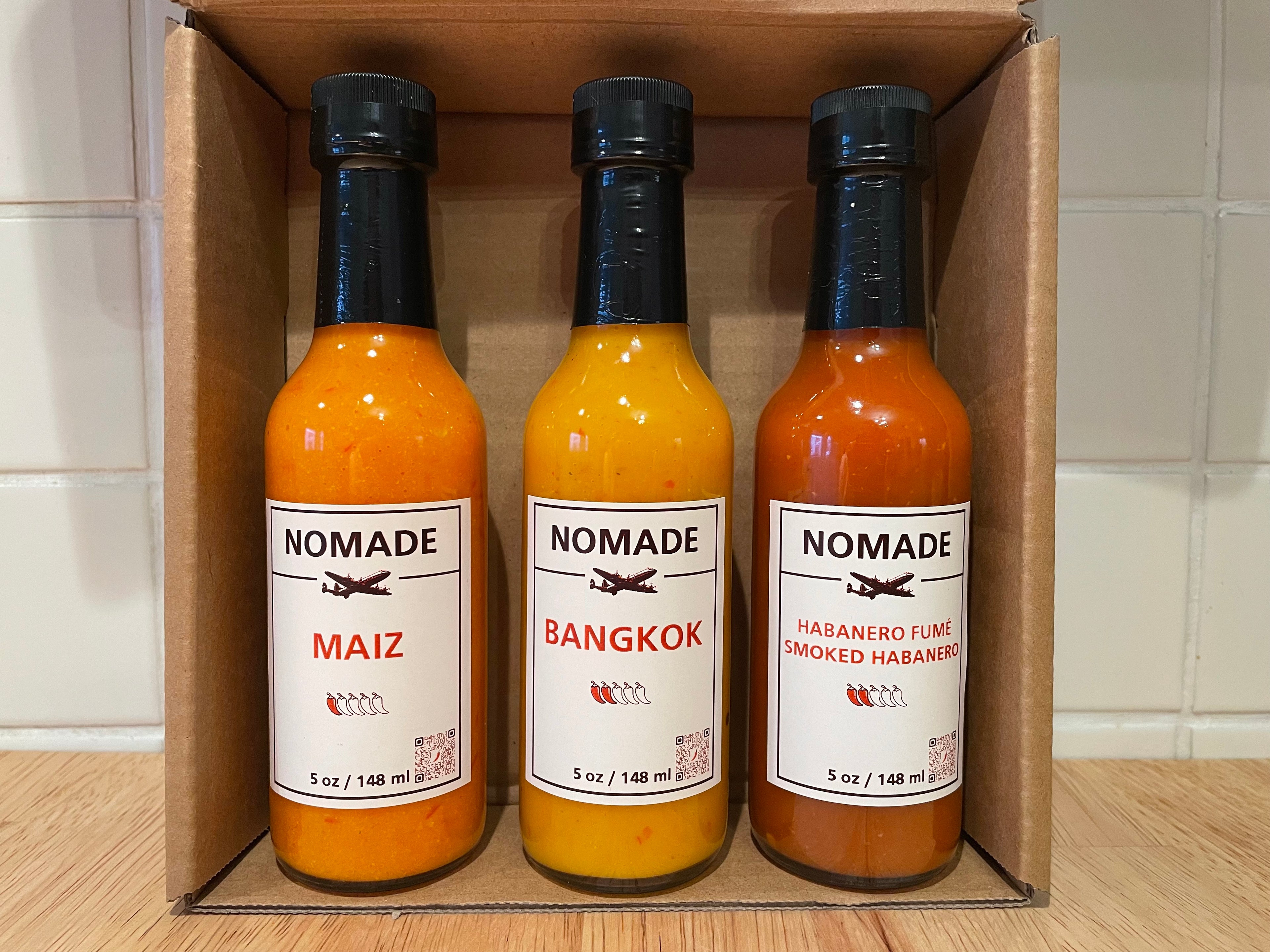 Three bottles of Nomade hot sauce in a cardboard box on a wooden surface.
